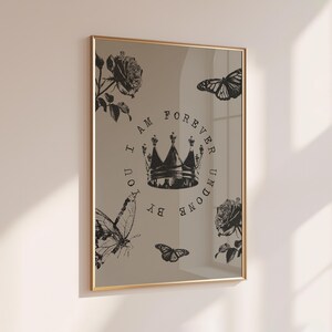May include: A framed print with a gold border. The print features a crown, roses, and butterflies in black and white. The text "I AM FOREVER UNDONE BY YOU" is printed around the crown. The print is displayed on a beige wall.