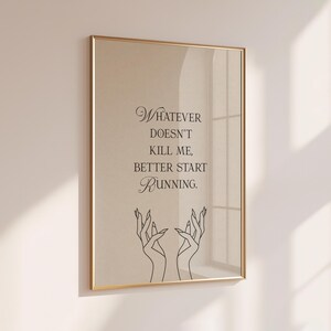 May include: A framed print with a gold-colored frame. The print features the quote "Whatever doesn't kill me, better start running" in black script. Below the text are two line-drawn hands with long fingers.