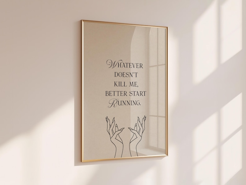 May include: A framed print with a gold-colored frame. The print features the quote "Whatever doesn't kill me, better start running" in black script. Below the text are two line-drawn hands with long fingers.