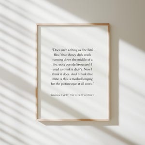 May include: A framed print with a light wood frame. The print features a quote from Donna Tartt's "The Secret History" in black text on a white background. The quote explores the concept of a 'fatal flaw' and a 'morbid longing'.