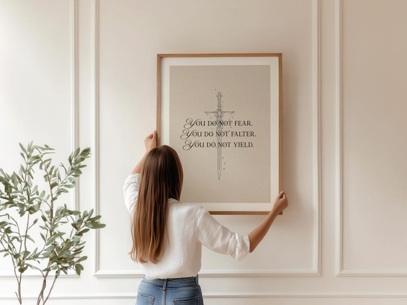 Może przedstawiać: Oprawiony wydruk z beżowym tłem i ilustracją miecza. Tekst brzmi "You do not fear. You do not falter. You do not yield." Rama jest w kolorze jasnobrązowym. Osoba trzyma oprawiony wydruk.