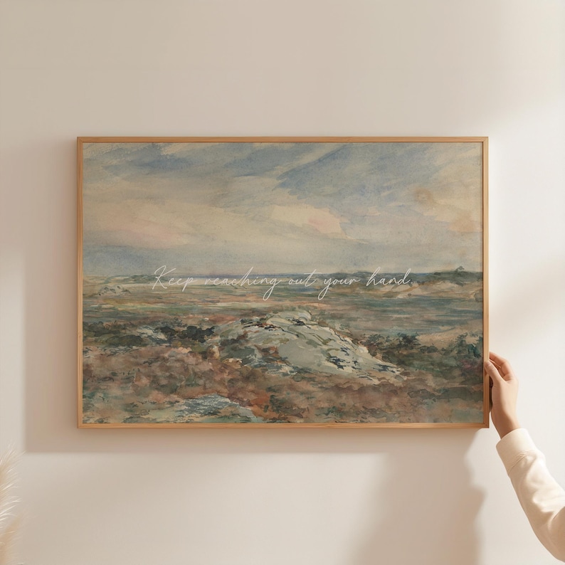 Op de afbeelding: Een ingelijste landschapsafdruk met een gedempt kleurenpalet van blauw, bruin en groen. Het kunstwerk toont een kustlandschap met een bewolkte lucht en rotsachtig terrein. De tekst "Keep reaching out your hand" staat over de afbeelding.