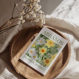 Puede incluir: Un plato de madera sostiene una postal con el texto "ABRAXOS FLOWER MARKET". La postal presenta una pintura de acuarela de narcisos amarillos y blancos. Flores blancas secas y sombras suaves completan la escena.