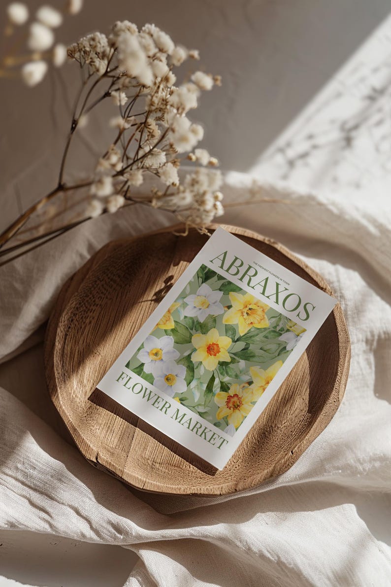 Puede incluir: Un plato de madera sostiene una postal con el texto "ABRAXOS FLOWER MARKET". La postal presenta una pintura de acuarela de narcisos amarillos y blancos. Flores blancas secas y sombras suaves completan la escena.