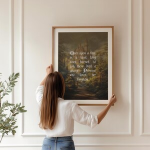 May include: A framed print with a fairytale scene of a castle in a forest, with the text "Once upon a time..." in elegant script. The print is held by a person in a white shirt and blue jeans.