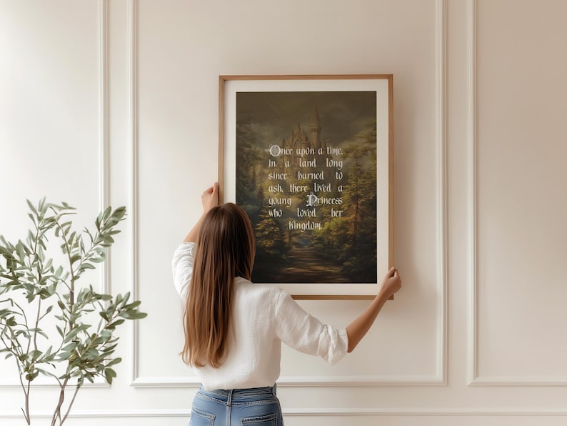 May include: A framed print with a fairytale scene of a castle in a forest, with the text "Once upon a time..." in elegant script. The print is held by a person in a white shirt and blue jeans.