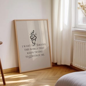 May include: A framed art print with a beige background and a wooden frame. The print features a black snake illustration with botanical accents and the text: "I WANT TO SURVIVE THIS WORLD THAT KEEPS TRYING TO DESTROY ME."