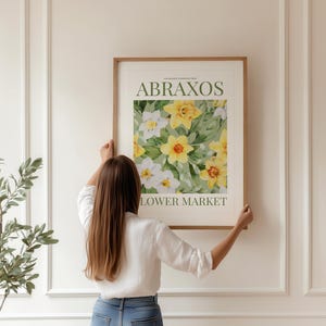 Puede incluir: Un cuadro enmarcado con un dise&ntilde;o floral de narcisos amarillos y blancos. El cuadro tiene el texto "ABRAXOS LOWER MARKET" en verde y negro. La obra de arte est&aacute; siendo sostenida por una persona con una camisa blanca y vaqueros azules.