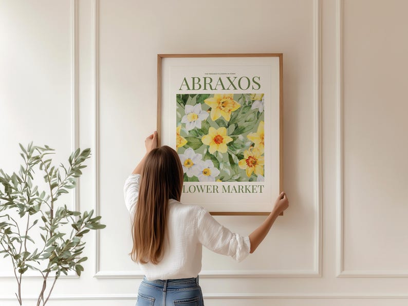 Puede incluir: Un cuadro enmarcado con un dise&ntilde;o floral de narcisos amarillos y blancos. El cuadro tiene el texto "ABRAXOS LOWER MARKET" en verde y negro. La obra de arte est&aacute; siendo sostenida por una persona con una camisa blanca y vaqueros azules.