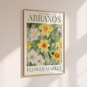Puede incluir: Un estampado floral enmarcado con narcisos amarillos y blancos y hojas verdes. El estampado tiene el texto "ABRAXOS FLOWER MARKET" en verde. El marco es dorado y cuelga de una pared blanca.