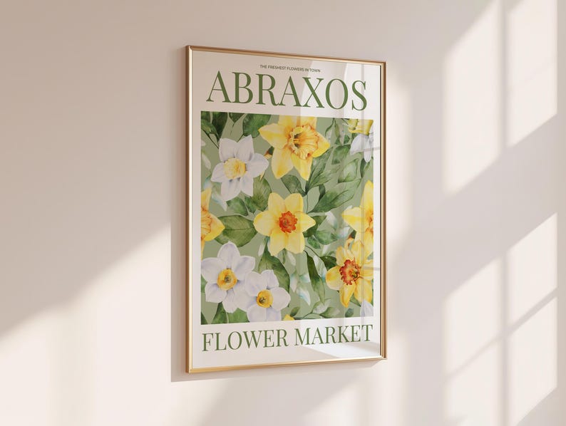 Puede incluir: Un estampado floral enmarcado con narcisos amarillos y blancos y hojas verdes. El estampado tiene el texto "ABRAXOS FLOWER MARKET" en verde. El marco es dorado y cuelga de una pared blanca.