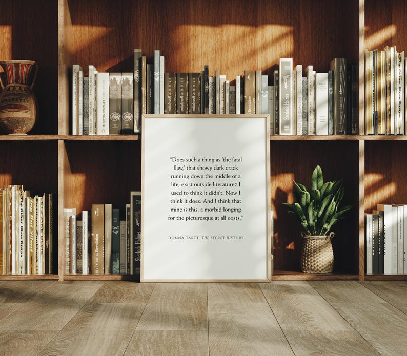May include: A framed print with text from Donna Tartt's "The Secret History" stands in front of a bookshelf filled with books. The print has a light wood frame and the text is in black. A small potted plant sits on the shelf.