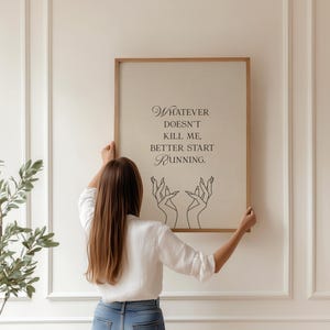 May include: A framed print with the quote "Whatever doesn't kill me, better start running." The artwork features elegant script and line art of hands. The frame is a light brown color, and the background is a neutral beige.
