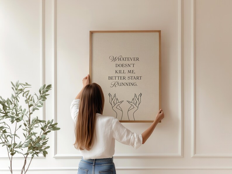May include: A framed print with the quote "Whatever doesn't kill me, better start running." The artwork features elegant script and line art of hands. The frame is a light brown color, and the background is a neutral beige.
