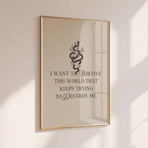 May include: A framed art print with a gold-colored frame. The print features a black snake intertwined with leaves above the quote, "I WANT TO SURVIVE THIS WORLD THAT KEEPS TRYING TO DESTROY ME." The background is a light beige color.