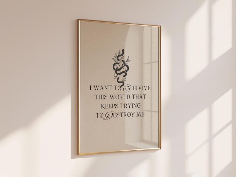 May include: A framed art print with a gold-colored frame. The print features a black snake intertwined with leaves above the quote, "I WANT TO SURVIVE THIS WORLD THAT KEEPS TRYING TO DESTROY ME." The background is a light beige color.