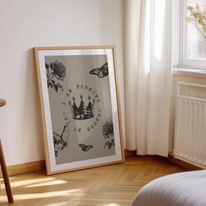 May include: A framed art print featuring a crown design with the text "I AM FOREVER UNDONE BY YOU". The artwork includes illustrations of roses and butterflies in black and white against a neutral background. The frame is a light wood color.
