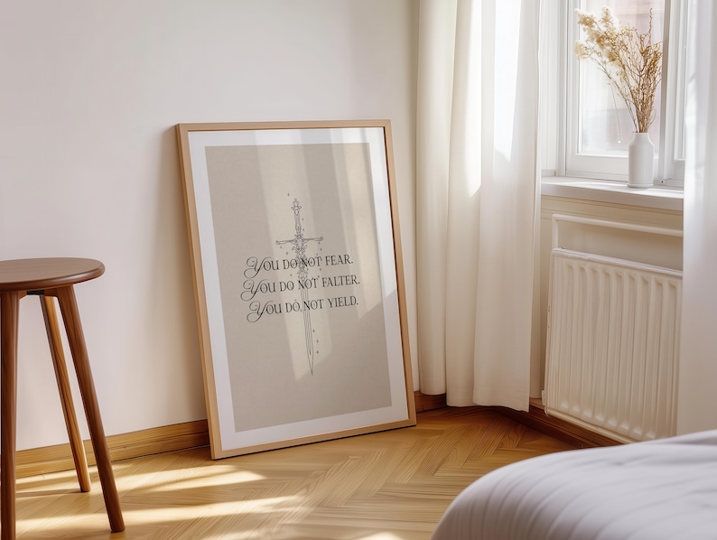 Może przedstawiać: Oprawiony wydruk z beżowym tłem i ilustracją miecza. Tekst brzmi: &bdquo;You do not fear. You do not falter. You do not yield.&rdquo; Rama jest w jasnobrązowym kolorze. Wydruk opiera się o ścianę.