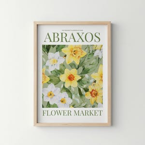 Puede incluir: Una impresi&oacute;n art&iacute;stica enmarcada con un dise&ntilde;o floral de narcisos amarillos y blancos y hojas verdes. El texto "ABRAXOS" y "FLOWER MARKET" est&aacute; impreso en verde. La impresi&oacute;n est&aacute; en un marco de madera clara.