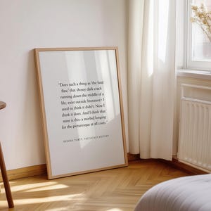 May include: A framed print with a quote from Donna Tartt's "The Secret History" in a minimalist style. The print is in a light wood frame, leaning against a white wall. A small wooden side table is in the foreground.