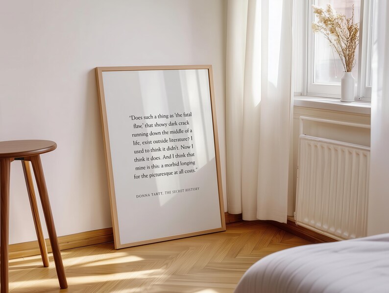 May include: A framed print with a quote from Donna Tartt's "The Secret History" in a minimalist style. The print is in a light wood frame, leaning against a white wall. A small wooden side table is in the foreground.