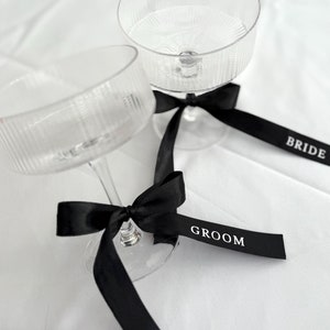 Puede incluir: Dos copas de champán de vidrio transparente con lazos de cinta negra atados alrededor de los tallos. Las cintas tienen las palabras "Bride" y "Groom" impresas en ellas.