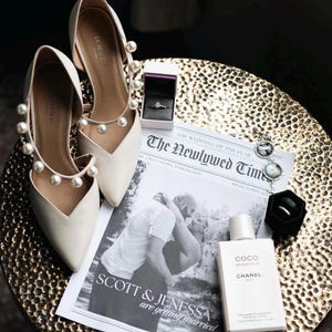 May include: Ivory-colored wedding shoes with pearl accents, a newspaper with the headline "The Newlywed Times", a ring box, and a bottle of Chanel Coco perfume are arranged on a gold-colored table. The newspaper features a black and white photo of a couple.