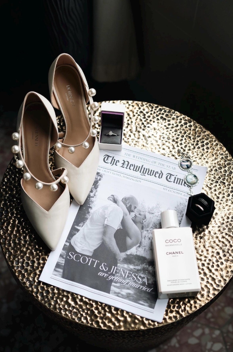 May include: Ivory-colored wedding shoes with pearl accents, a newspaper with the headline "The Newlywed Times", a ring box, and a bottle of Chanel Coco perfume are arranged on a gold-colored table. The newspaper features a black and white photo of a couple.
