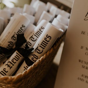 May include: A woven basket filled with rolled-up newspapers tied with black ribbons. The newspapers are labeled "The Newlywed Times" and "The Year". A sign with elegant script is visible in the background.