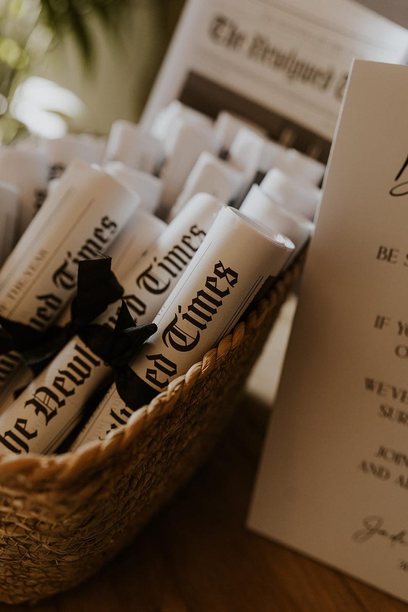 May include: A woven basket filled with rolled-up newspapers tied with black ribbons. The newspapers are labeled "The Newlywed Times" and "The Year". A sign with elegant script is visible in the background.