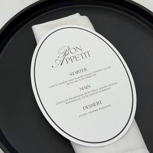 Puede incluir: Una bandeja negra con una tarjeta de menú ovalada blanca y una servilleta blanca doblada. La tarjeta del menú dice "Bon Appétit" y enumera un entrante, un plato principal y un postre. El menú incluye queso de cabra, ñoquis y pudín de caramelo.