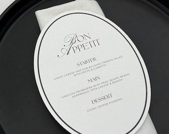 May include: A black serving tray with a white oval menu card and a folded white napkin. The menu card reads "Bon Appetit" and lists a starter, main course, and dessert. The menu includes goat cheese, gnocchi, and sticky toffee pudding.