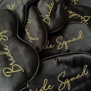 May include: A close-up of six black satin sleep masks with gold lettering that reads "Bride Squad".