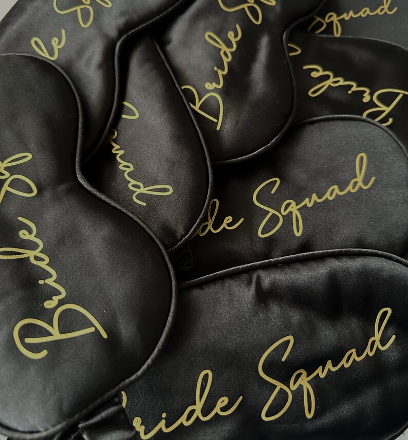 May include: A close-up of six black satin sleep masks with gold lettering that reads "Bride Squad".