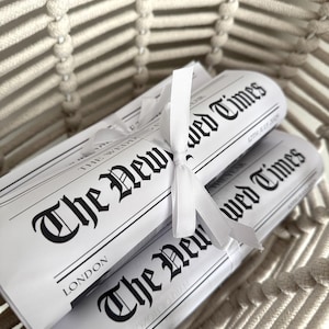 May include: Two rolled newspapers tied with a white ribbon, nestled in a woven basket. The newspapers are titled "The New Wed Times" with the date "12th July 2009" and the city "London" printed on them. The basket is made of a light beige rope.