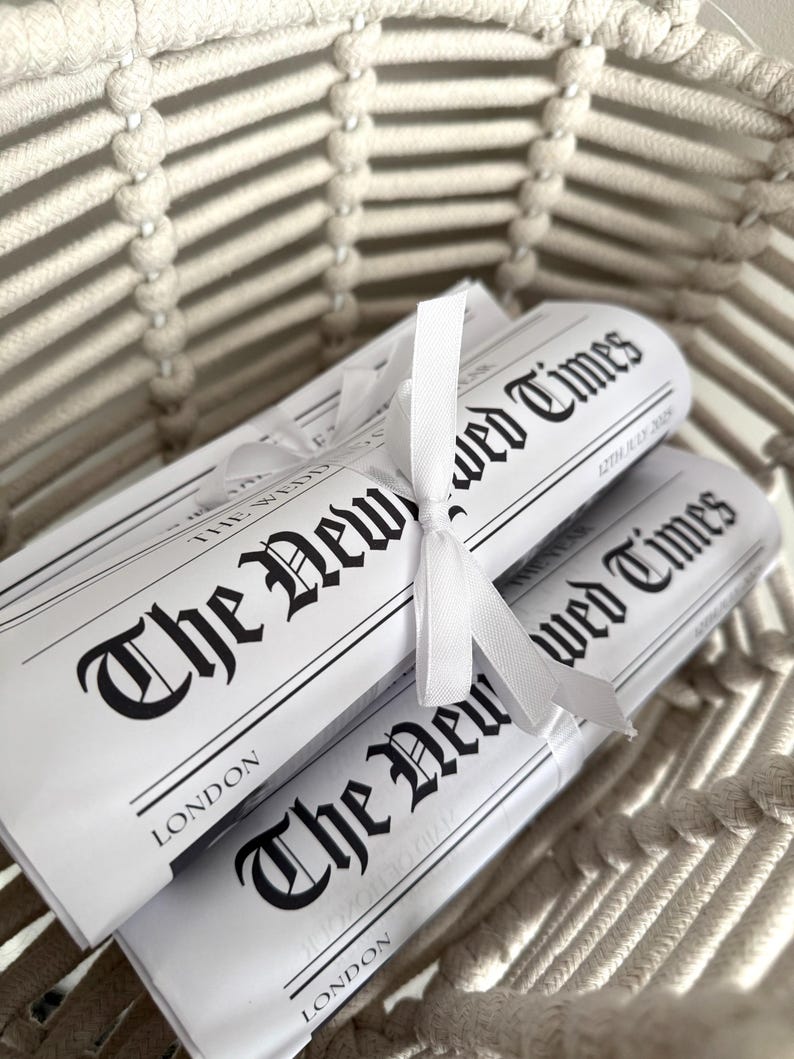 May include: Two rolled newspapers tied with a white ribbon, nestled in a woven basket. The newspapers are titled "The New Wed Times" with the date "12th July 2009" and the city "London" printed on them. The basket is made of a light beige rope.