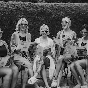 May include: Black and white photo of five people wearing heart-shaped sunglasses and robes, seated outdoors. Each person holds a newspaper. The scene suggests a relaxed, social gathering.