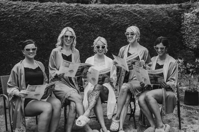 May include: Black and white photo of five people wearing heart-shaped sunglasses and robes, seated outdoors. Each person holds a newspaper. The scene suggests a relaxed, social gathering.