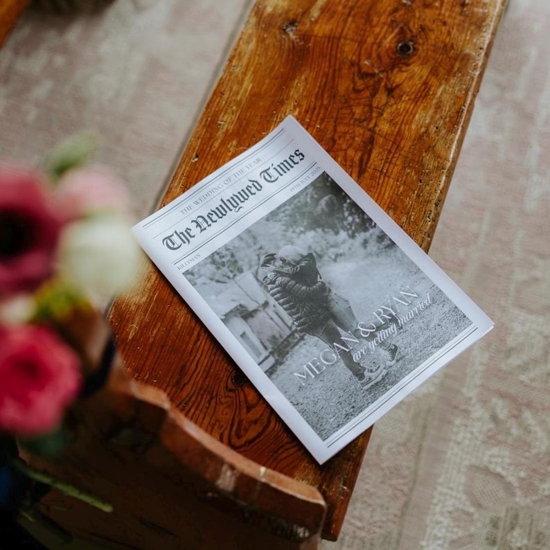 May include: A newspaper titled "The Newlywed Times" rests on a wooden bench. The black and white image on the front page shows a couple embracing. The headline reads "MEGAN & RYAN are getting married."