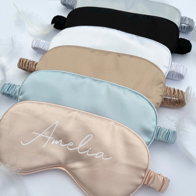May include: A set of six satin sleep masks in various colors, including black, white, beige, light blue, and a light pink mask with the name "Amelia" embroidered in white.