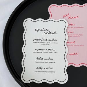 May include: Two decorative cards with scalloped edges sit on a black tray. One card lists "signature cocktails" with martini recipes. The other card, in pink, says "girl dinner" with menu items. The text is in black and red.