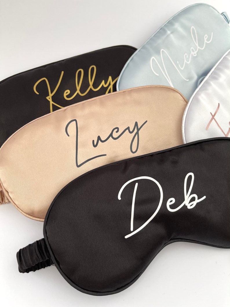 May include: Four personalized sleep masks in different colors. The masks are made of satin and have names written on them in gold and white lettering. The names are Kelly, Lucy, Nicole, and Deb.