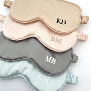 May include: Four satin sleep masks in different colors, stacked on top of each other. The top mask is beige with black embroidered initials "KD". The second mask is pink with white embroidered initials "RM". The third mask is gray with white embroidered initials "MB". The bottom mask is light blue with white embroidered initials "RM".