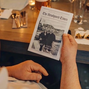 May include: A person holding a black and white newspaper titled "The Newlywed Times." The newspaper features a photograph of a couple and the names "Claire & Andi." The background includes a wooden table, glassware, and silverware.