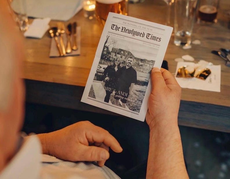 May include: A person holding a black and white newspaper titled "The Newlywed Times." The newspaper features a photograph of a couple and the names "Claire & Andi." The background includes a wooden table, glassware, and silverware.