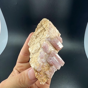 May include: A raw mineral specimen featuring clusters of translucent, pale pink, prismatic crystals embedded within a rough, beige, and tan-colored matrix. The crystalline structure displays vertical striations and a glassy luster throughout the formation.