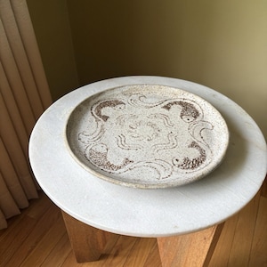 May include: A round, speckled ceramic plate with a fish design. The plate is off-white with brown detailing and sits on a white marble table. The fish design is repeated four times around the plate's edge.