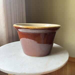 May include: A brown ceramic planter with a wide, slightly flared rim. The planter has a two-tone design, with a lighter tan color at the rim and a darker brown body. The planter is sitting on a white marble surface.