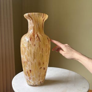 May include: A tall glass vase featuring a swirling pattern of cream, beige, and dark brown hues. The vessel has a flared rim, a slender neck, and a rounded body with a smooth, glossy finish that reflects light across its mottled, organic surface.
