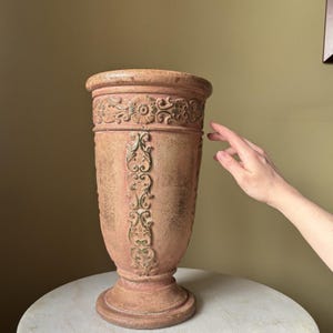 May include: A tall, terracotta-colored decorative vase with ornate floral and scrollwork detailing. The vase has a wide top, a slender body, and a pedestal base. The surface has a textured, aged appearance, suggesting a vintage or antique style.